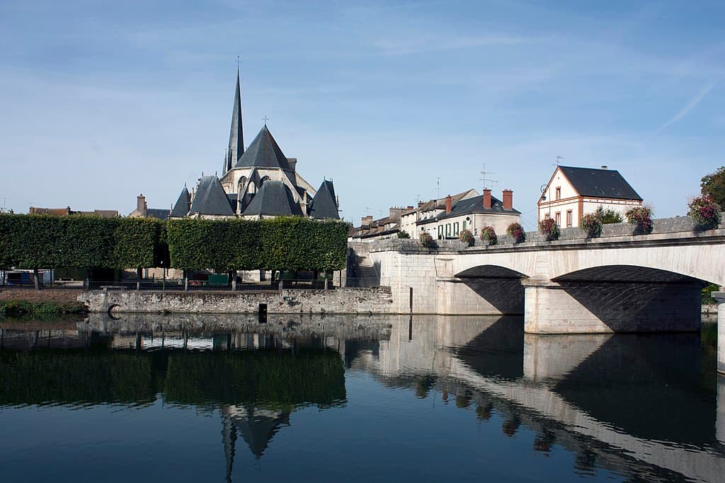 Église au bord de la rivière avec un pont en pierre et des arbres taillés à Nemours, sous un ciel bleu clair.