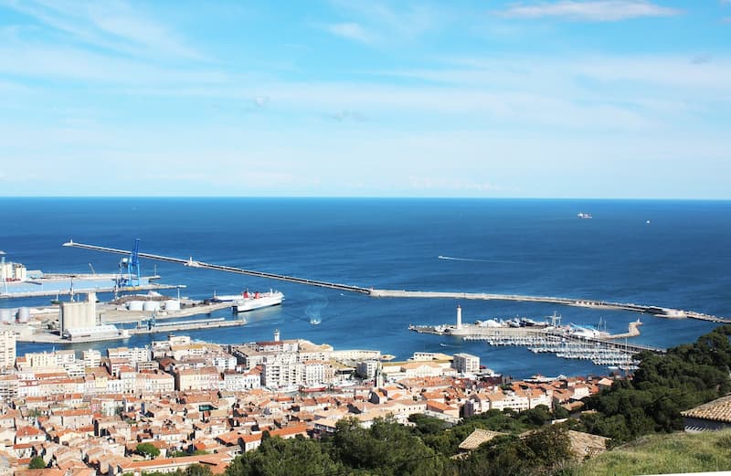 Vue aérienne du port de Sète montrant le port industriel, le phare, la marina et la ville historique avec ses toits orangés, bordée par la Méditerranée