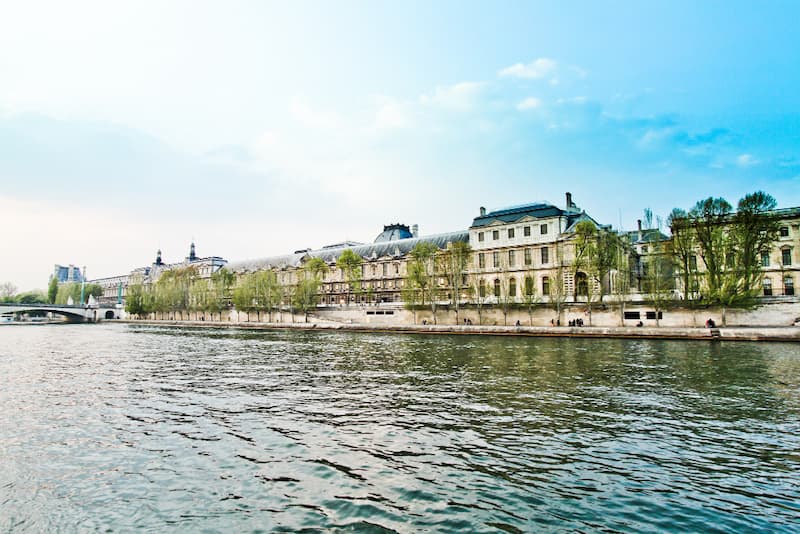 Vue panoramique du Musée du Louvre depuis la Seine à Paris, montrant la façade historique du palais et les quais bordés d'arbres par une journée ensoleillée